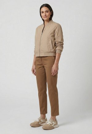 Kvinde stående iført en beige jakke med lynlås, brune bukser og beige sneakers mod en ensartet lysegrå baggrund.