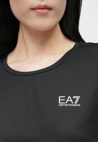 Fekete rövid ujjú póló kerek nyakkivágással, bal oldalon fehér EA7 Emporio Armani logóval. Puha, sima tapintású anyag.