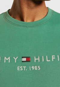 Groene sweatshirt met een ronde hals, geborduurd logo in het wit en een kleurrijk embleem met rode, witte en zwarte accenten.