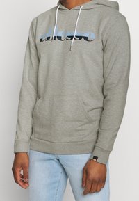 Sudadera ligera gris con capucha ajustable, bolsillo frontal y un gran logo en azul y negro. Puños y dobladillo acanalados con una etiqueta de la marca.