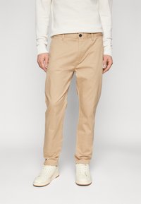 Chinos beige en coton, avec une coupe droite, des poches avant et une fermeture à bouton, associés à des baskets blanches.