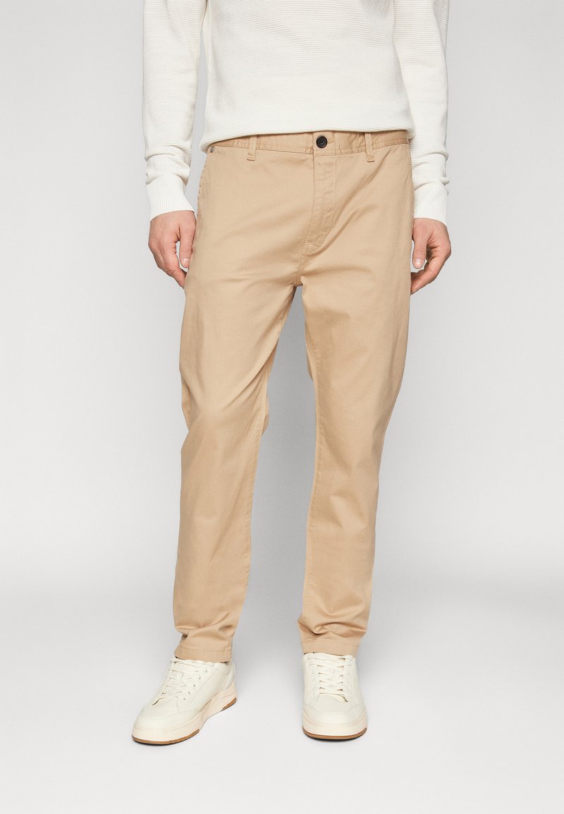 Chinos beige en coton, avec une coupe droite, des poches avant et une fermeture à bouton, associés à des baskets blanches.