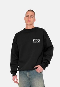 Schwarzer Hoodie mit lockerer Passform und gerippten Bündchen. Mit einem aufgenähten Brustpatch, das ein strukturiertes Muster aufweist. Kombiniert mit hellblauem Denim.