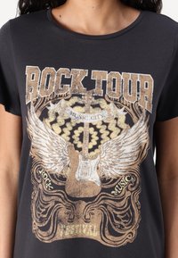 T-shirt noir avec un imprimé graphique représentant une guitare, des ailes et le texte "ROCK TOUR" dans des tons dorés et argentés. Manches courtes, col rond.