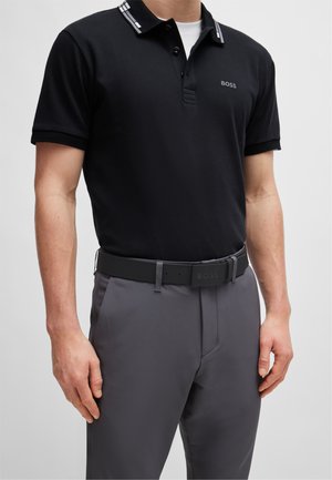 Polo noir avec un col comportant des rayures blanches et bleues, associé à un pantalon gris et une ceinture noire avec logo. Tissu lisse.