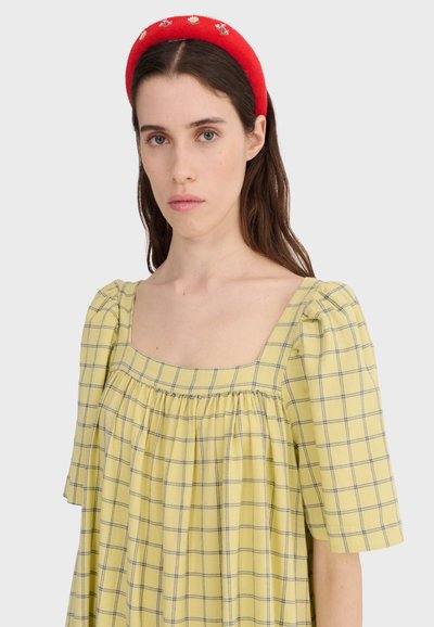 Femme aux longs cheveux bruns portant une robe à carreaux jaune avec des manches bouffantes et un bandeau rouge brodé de motifs floraux.