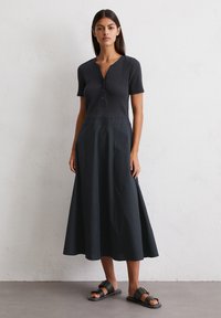 Model stands confidently in a dark, fitted top with buttons, paired with a flowing black skirt and flat sandals, against a neutral background.