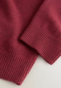 Burgundská pletenina s hladkou texturou, s žebrovanými manžetami a měkkým, mírně chlupatým povrchem. Minimalistický design bez vzorů.