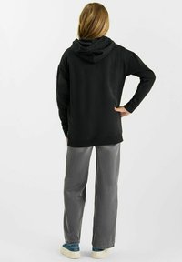 Persoon met lang haar staat met de rug naar de kijker, gekleed in een zwarte hoodie, grijze broek en blauwe sneakers, voor een witte achtergrond.