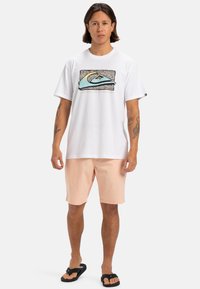 Camiseta blanca de manga corta con un gráfico azul y negro en el frente, combinada con pantalones cortos rosa claro y chanclas negras.