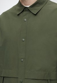 Homme portant une chemise boutonnée vert olive avec un col, présentant cinq boutons mats visibles sur le devant.