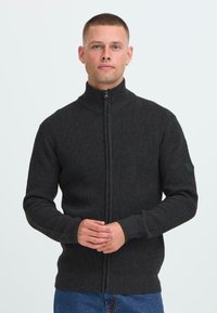Uomo con i capelli corti che indossa un maglione in maglia grigio scuro con zip, in piedi con le mani incrociate, su uno sfondo chiaro e semplice.