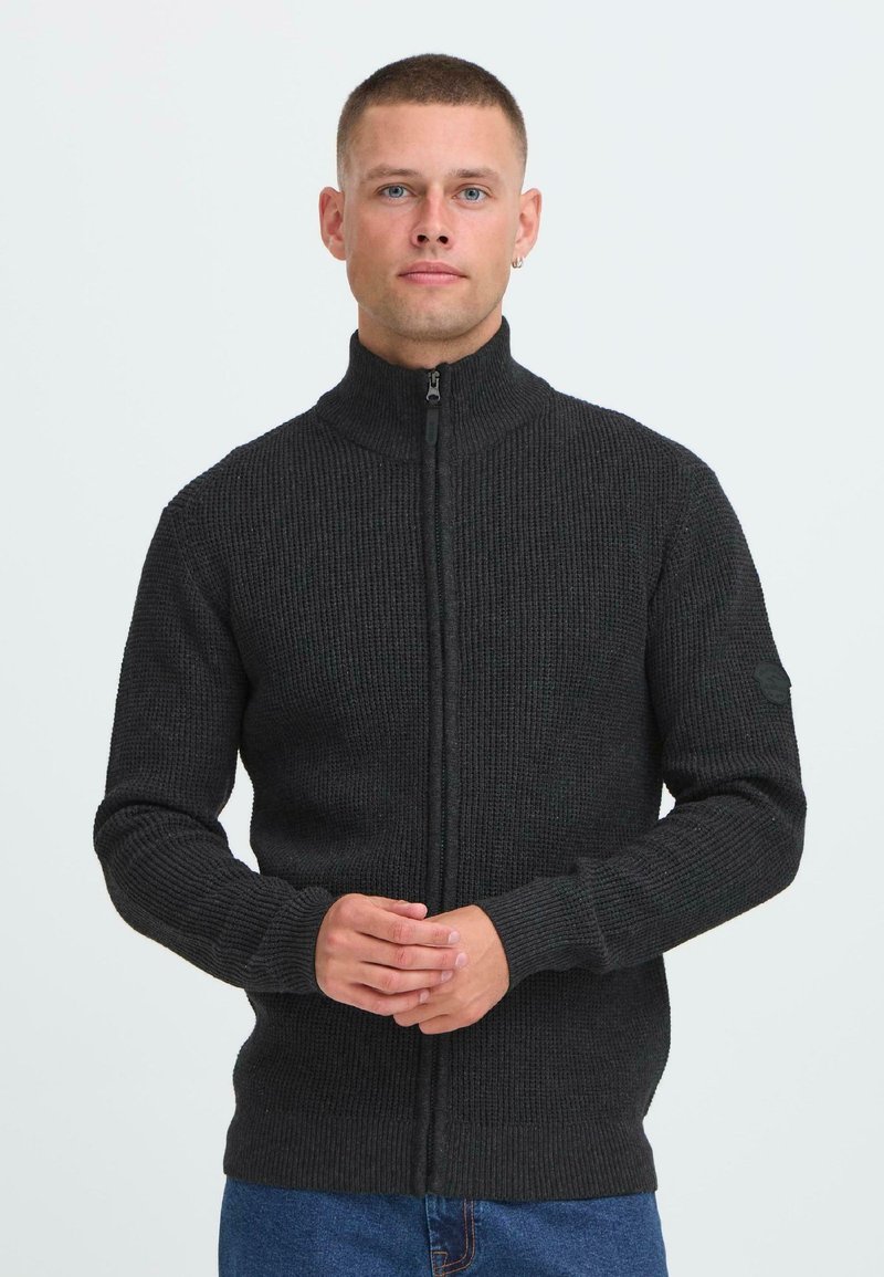 Uomo con i capelli corti che indossa un maglione in maglia grigio scuro con zip, in piedi con le mani incrociate, su uno sfondo chiaro e semplice.