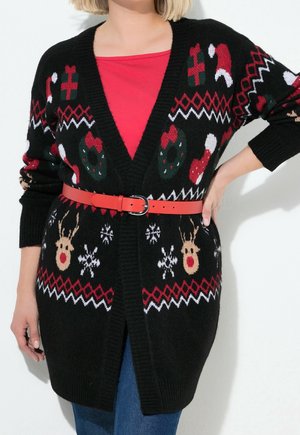 Femme porte un cardigan de Noël noir avec des rennes, des couronnes, des cadeaux et des motifs de flocons de neige, ceinturé sur une chemise rouge et un jean bleu.