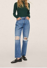 Frau mit grünem, geripptem Langarmoberteil, hochtaillierten, zerrissenen blauen Jeans und schwarzen Mokkasins, die mit den Händen in den Taschen steht.