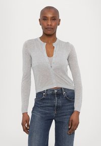 Světle šedý zkrácený cardigan vyrobený z měkkého pleteného materiálu. Má kulatý výstřih a zapínání na knoflíky, doplněný tmavě modrými džíny.