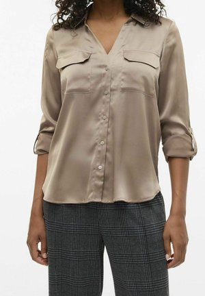 Femme portant un chemisier beige boutonné avec manches retroussées et pantalon gris à carreaux, debout avec les bras détendus le long du corps.