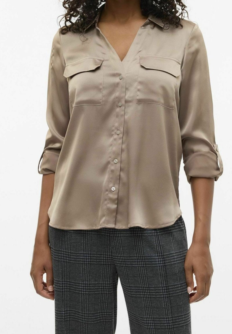Femme portant un chemisier beige boutonné avec manches retroussées et pantalon gris à carreaux, debout avec les bras détendus le long du corps.