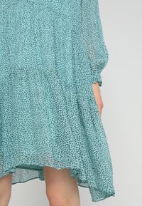 Plynná, patrová šaty v světle tyrkysové barvě s leopardím vzorem. Průhledná tkanina s elastickými manžetami a lemováním, které je vpředu kratší.