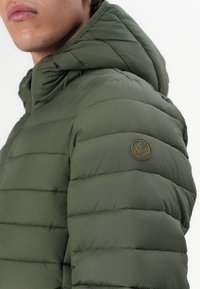Green padded jacket with a hood, featuring horizontal stitching, a textured finish, and a circular logo patch on the shoulder.