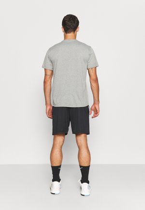 Homme debout de dos portant une chemise grise à manches courtes, un short noir, des chaussettes noires et des chaussures de sport blanches sur un fond uni.