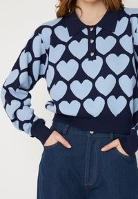 Pulôver de malha azul-marinho com um padrão de corações azul-claro, apresentando um decote com gola e botões em forma de coração. Combina com jeans de algodão escuro.