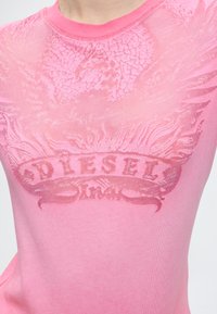 Κοντινό πλάνο σε ένα ροζ t-shirt Diesel με ανάγλυφο σχέδιο φλόγας και πανό που φέρει το όνομα της μάρκας στο στήθος.