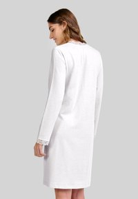 Mujer vestida con un vestido blanco de manga larga hasta la rodilla con encaje en los puños y el escote, de pie frente a un fondo gris liso.