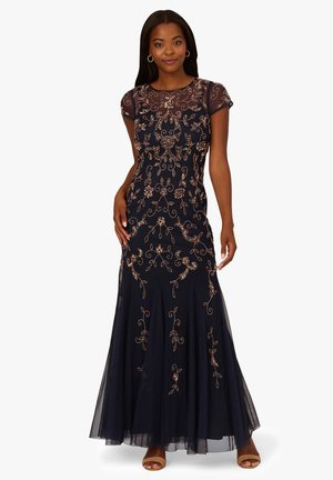 Vrouw in een marineblauwe jurk tot aan de grond met korte mouwen en verfijnde gouden bloemenborduursels, gecombineerd met bruine open-toe hakken.