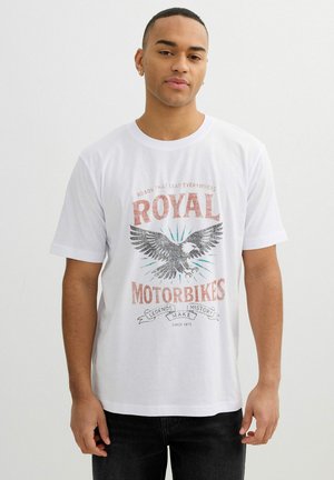 Joven con una camiseta blanca con un gráfico vintage de un águila y el texto "Royal Motorbikes Legends History Make Since 1975."