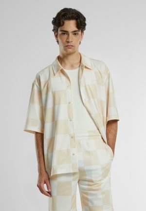 Camisa de botonadura corta en un patrón de cuadros en beige y blanco. Textura suave, corte relajado, con un sutil detalle del logo en el pecho.