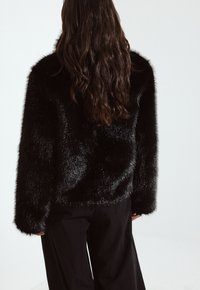 Melnas mākslīgās kažokādas jaka ar mīkstu tekstūru, ar kastes griezumu un platām piedurknēm. Uzvilkta virs melniem biksēm, uzrādot spīguļojošu apdari.