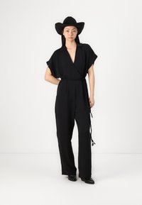 Svart jumpsuit med dyp V-hals, korte ermer og innsiddet midje med knyttet belte. Brukt med en svart bredbremmet hatt og svarte sko.