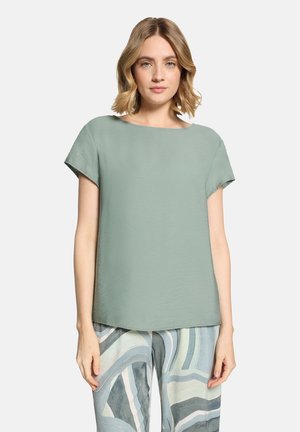 Frau mit schulterlangem blondem Haar, die ein lockeres, kurzärmeliges Oberteil in Minzgrün und abstrakt gemusterte Hosen in Blau- und Grautönen trägt.