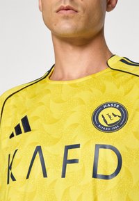 Yellow jersey with swirling pattern, featuring black stripes and logo. Material is lightweight and smooth with a crew neck design.
