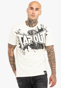 Bijela majica s velikim crnim "TAPOUT" grafičkim otisakom u stilu prskanja. Kratki rukavi, okrugli izrez i oznaka na donjoj strani.