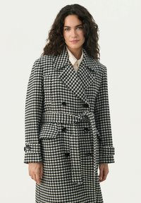 Double-breasted houndstooth coat in black and white, featuring a belted waist, large lapels, and button accents on the sleeves.