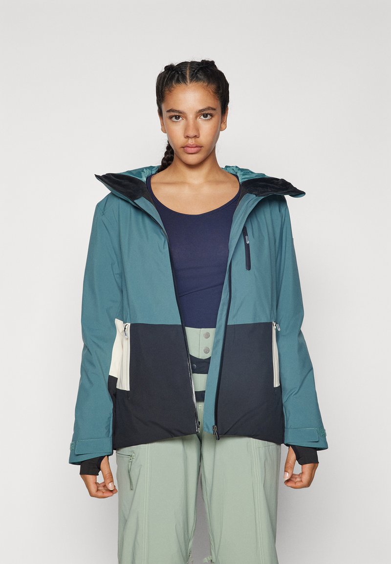 Jacke mit einem teakfarbenen oberen Abschnitt, einem schwarzen unteren Abschnitt und cremefarbenen Akzenten. Verfügt über eine Kapuze, Reißverschlusstaschen und eine glatte, wasserdichte Textur.