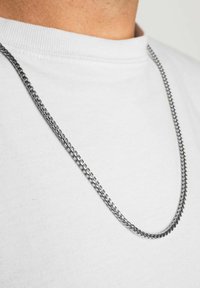 Colier din argint cu un design răsucit, atârnă deasupra unei cămăși albe cu guler rotund, evidențiind o suprafață netedă și un finisaj reflexiv.