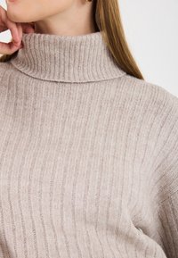 Pull à col roulé côtelé beige clair avec une texture douce et un col haut, présentant une coupe ajustée et des rayures verticales.