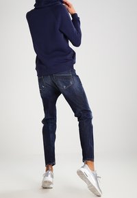 En kvinna står med ryggen mot kameran, klädd i en marinblå huvjacka, mörka jeans och silverfärgade sneakers, i en minimalistisk miljö.