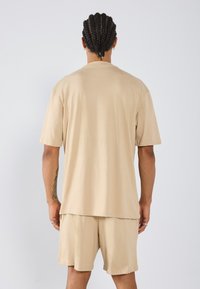 Beige rövid ujjú ing laza szabással, azonos színű bézs rövidnadrággal párosítva. Puha pamut anyag, sima textúrával, egyszerű dizájn.