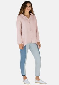 Eine Frau trägt ein lockeres, pinkes Langarmshirt, zweifarbige, blaue Skinny-Jeans, weiße Slipper und goldenen Schmuck und steht vor einem weißen Hintergrund.