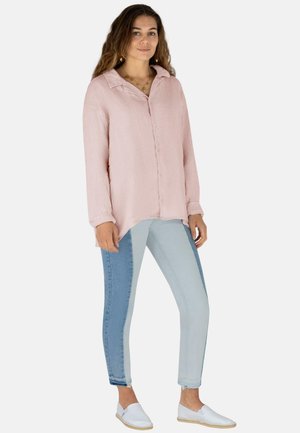 Eine Frau trägt ein lockeres, pinkes Langarmshirt, zweifarbige, blaue Skinny-Jeans, weiße Slipper und goldenen Schmuck und steht vor einem weißen Hintergrund.