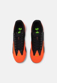 Baskets de sport noires et orange avec une tige texturée, comportant des accents verts et une semelle en caoutchouc bleue. Design à lacets avec un branding marqué.