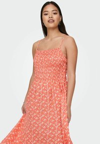 Koralinė florinė suknelė su smulkinta liemene, plonomis petnešėlėmis ir plačiu sijonu. Pasižymi baltais gėlių raštais ant ryškios fono.