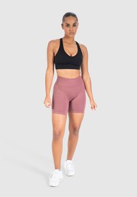 Sujetador deportivo negro y pantalones cortos de talle alto en color rosa marrón, hechos de una tela elástica. La modelo está de pie en zapatillas blancas frente a un fondo gris.