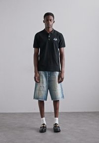 Svart polo-skjorte med logo, blå slitte denimshorts, hvite sokker og svarte sko. Modellen står mot en enkel bakgrunn.