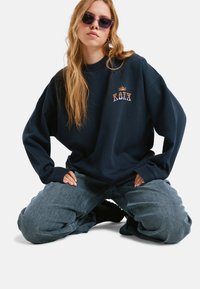 Granatowy, oversize'owy sweter z haftowanym logo "ROXY", zestawiony z luźnymi szarymi dżinsami. Okulary przeciwsłoneczne dodają nowoczesnego charakteru.
