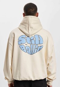 Crème kleurige katoenen hoodie met een ruime capuchon, oversized pasvorm en grote blauwe "Still Ballin"-graphic op de achterkant. Zachte textuur en geribde manchetten.